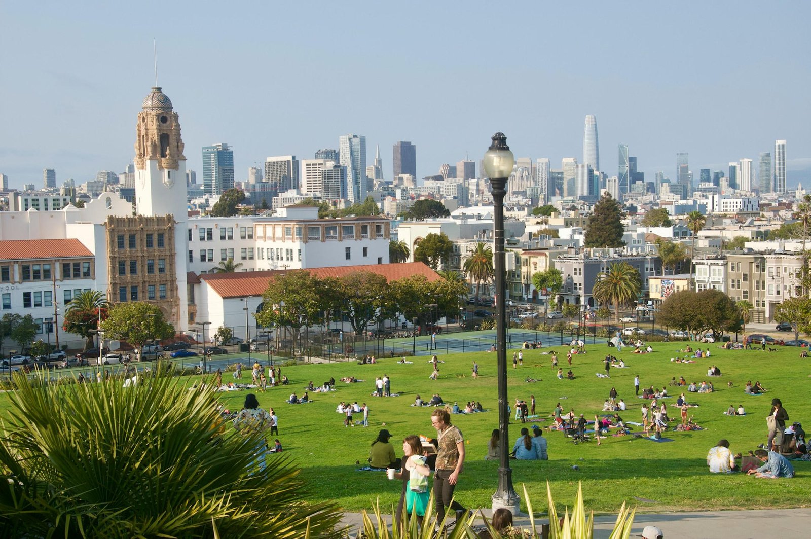 Lush green park filled with people enjoying sunny day, city skyline in background